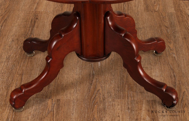 May include: Close-up of the base of a mahogany table with three legs. Each leg has a carved design and ends in a rounded foot with a brass caster.