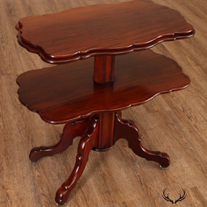 May include: A two-tiered mahogany table with a scalloped edge on each tier. The table has a turned pedestal base with four legs that are carved with a scroll design. The table is in good condition and has a rich, dark brown finish.