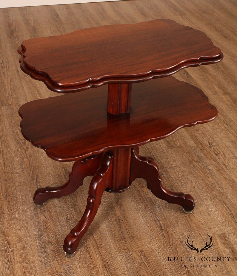 May include: A two-tiered mahogany table with a scalloped edge on each tier. The table has a turned pedestal base with four legs that are carved with a scroll design. The table is in good condition and has a rich, dark brown finish.