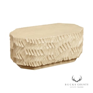 May include: A large, oval-shaped coffee table with a beige, textured surface. The table has a geometric pattern carved into the sides and a slightly raised base.