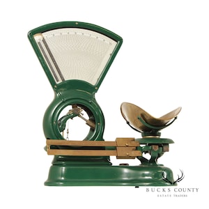 May include: A vintage green and brass scale with a circular dial and a large, brass weighing pan. The scale has a brass arm with two weights and a hanging weight.