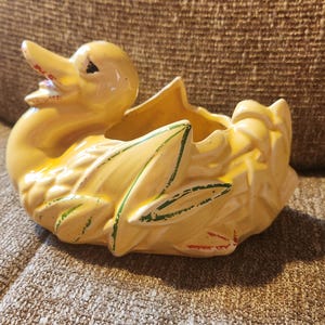 Op de afbeelding: Een gele keramische eendenplanter met groene en rode accenten. De eend heeft een gedetailleerd ontwerp, met een getextureerd lichaam en een brede opening voor planten. Een decoratief item voor woondecoratie.
