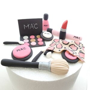 May include: A white cake decorated with a makeup theme. The cake is decorated with a pink and black makeup palette, a pink lipstick, a pink nail polish bottle, a black makeup brush, and a pink and black compact with the word "M".A.C." written on it.