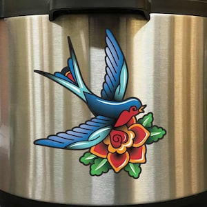 May include: A stainless steel Instant Pot with a blue and red swallow tattoo sticker on the side. The swallow is flying with its wings spread and is holding a red rose in its beak.