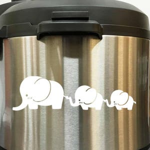 May include: Stainless steel electric pressure cooker with a black lid and a white elephant family design on the front.