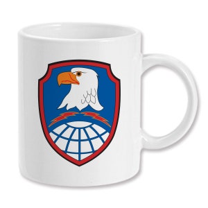 May include: White ceramic coffee mug with a red, white, and blue eagle emblem. The eagle is holding a globe with lightning bolts. The emblem is surrounded by a red border.