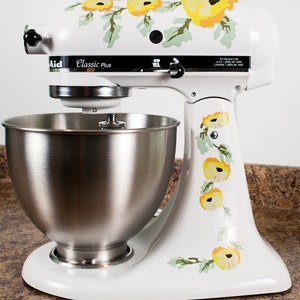 May include: White stand mixer with a silver bowl and a floral design featuring yellow flowers and green leaves. The mixer has the text "Aid Classic Plus" on the side.