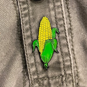 May include: A yellow and green enamel pin of a corn cob with husks.