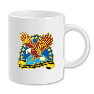 May include: White ceramic coffee mug with a graphic of a red-tailed hawk with a yellow beak and talons, flying over a blue globe with white stars. The globe is surrounded by a yellow banner with the text "SECURE THE NIGHT GROUND".