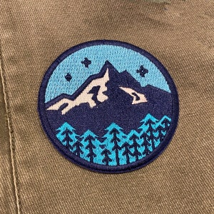 May include: A blue and white embroidered patch featuring a mountain range and trees. The mountain range is dark blue with white snow peaks. The trees are a light blue colour.