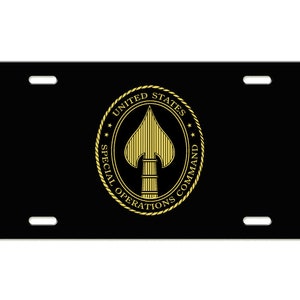 May include: Black license plate featuring a gold emblem of a spade within a circle, with the text "UNITED STATES SPECIAL OPERATIONS COMMAND". The plate has four mounting holes.