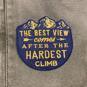 May include: A blue and gold embroidered patch with a mountain range design and the text "THE BEST VIEW comes AFTER THE HARDEST CLIMB".