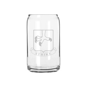 May include: A clear glass can-shaped drinking glass. The glass features a gray emblem of a bird's talon within a shield, with the word "STRIKE" on a banner below. The glass is designed for beverages.