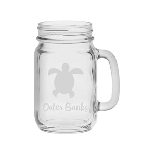 Puede incluir: Una taza de vidrio transparente en forma de tarro de albañil con asa. La taza tiene una silueta blanca de una tortuga marina y el texto "Outer Banks" grabado en el lateral.
