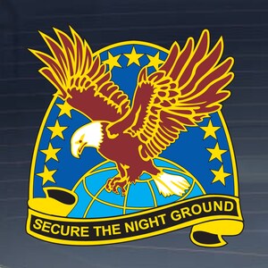 May include: A blue and yellow emblem with a red-brown eagle with white head and wings spread, holding a globe with a blue and white design. The text "SECURE THE NIGHT GROUND" is on a yellow banner.