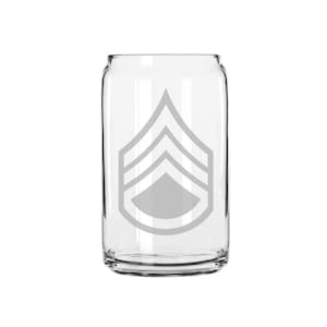 May include: A clear glass can-shaped drinking glass with a gray military rank insignia. The insignia features a chevron design with three stripes and a central emblem. The glass is empty and sits against a white background.