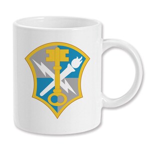 May include: White ceramic coffee mug with a gold, blue, and gray military insignia featuring a key, torch, and lightning bolt.