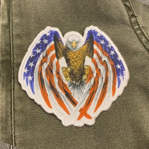 May include: A white, red, and blue embroidered patch featuring an eagle with its wings spread wide. The eagle's wings are formed by the American flag.