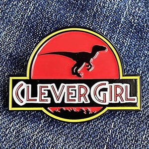 May include: A black and red enamel pin with a silhouette of a dinosaur in a red circle and the text "Clever Girl" in white with a black outline.