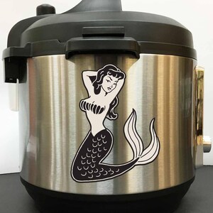 May include: A stainless steel electric pressure cooker with a black lid and a black and white mermaid sticker on the front.
