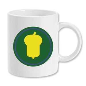 Può includere: Tazza da caffè in ceramica bianca con un cerchio verde che contiene una silhouette di una ghianda gialla. Il cerchio ha un sottile contorno blu.