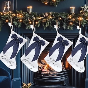 Puede incluir: Cuatro calcetines de Navidad blancos con lazos azul marino y nombres personalizados: Papá, Harry, Storie y Abuela G. Los calcetines están colgados en una repisa de chimenea con una chimenea encendida debajo.
