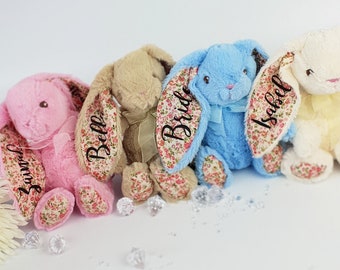 May include: Four plush stuffed bunnies in pink, tan, blue, and cream. Each bunny features long ears and foot pads with a floral pattern. The ears are personalized with black script text reading "Jauvel," "Belle," "Bride," and "Isabel."