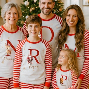 Puede incluir: Un grupo de personas con pijamas navideños a juego a rayas rojas y blancas. Los pijamas tienen una parte superior blanca con una letra y un motivo festivo, como un reno o Papá Noel. La familia sonríe frente a un árbol de Navidad.
