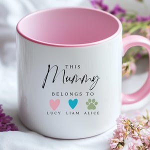 May include: White ceramic mug with a pink rim and handle. The mug has black text that reads "This Mummy Belongs To" with three icons below: a pink heart, a blue heart, and a paw print. The icons are labeled "Lucy", "Liam", and "Alice".