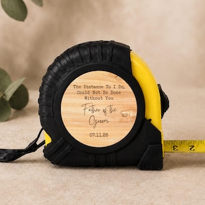 May include: A yellow and black retractable tape measure with a wood-grain circular insert. The text reads: The Distance To I Do, Could Not Be Done Without You, Father of the Groom, 07.11.26. A yellow metal tape extends to show markings in inches.