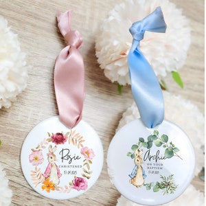 Può includere: Due ornamenti rotondi in ceramica con un design di Peter Rabbit. L'ornamento a sinistra è rosa con accenti floreali e dice "Rosie Christened 17.04.2023". L'ornamento a destra è blu con foglie verdi e dice "Archie On Your Baptism 17.04.2023".