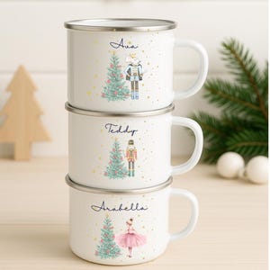 Puede incluir: Tazas de esmalte blanco apiladas, cada una con borde y asa plateados. Las tazas presentan nombres personalizados, ilustraciones de árboles de Navidad y personajes festivos como un cascanueces y una bailarina. Acentos de estrellas doradas.