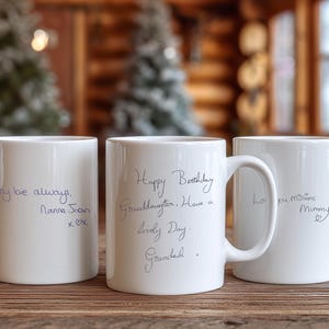 May include: Three white ceramic mugs with handwritten messages. The mugs have messages like "my love always" and "Happy Birthday Granddaughter". The mugs are on a wooden surface.
