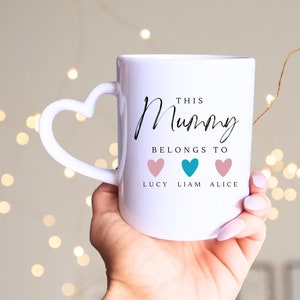May include: White ceramic mug with a heart-shaped handle. The mug has black text that reads "This Mummy Belongs To" with three hearts underneath. The hearts are pink, teal, and pink, with the names "Lucy", "Liam", and "Alice" written underneath.