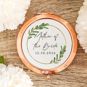 May include: A round rose gold compact mirror featuring a white insert with a gold circular frame and green leaf illustrations. The text reads Mother of the Bride and 12.10.2026 in a black script and serif font, centered within the botanical design.
