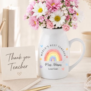 May include: A white ceramic pitcher filled with a bouquet of pink and white daisies. The pitcher features a watercolor rainbow graphic with the text "WORLD'S BEST TEACHER," "Miss Adams," and "Love Evan." A card reads "Thank you Teacher" with a heart.