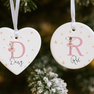 May include: Two white ceramic Christmas ornaments. One is heart-shaped with a pink letter "D" and the name "Daisy". The other is round with a pink letter "R" and the name "Rui". Both feature a ballerina illustration and star accents.