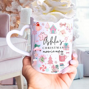 Op de afbeelding: Een witte keramische mok met een hartvormig handvat, gevuld met slagroom. De mok is versierd met kerstthema-illustraties in roze en groen, waaronder peperkoekenmannetjes, een auto en een kerstboom. De tekst "Ashlie's Christmas movie mug" staat ook op de mok.