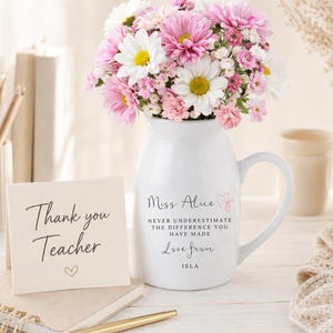 Puede incluir: Una jarra de cerámica blanca llena con un ramo de margaritas rosas y blancas y paniculata. La jarra presenta el texto "Miss Alice", "NEVER UNDERESTIMATE THE DIFFERENCE YOU HAVE MADE", "Love from" e "ISLA". Una tarjeta dice "Thank you Teacher".