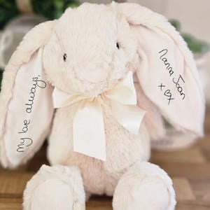 Könnte beinhalten: Ein cremefarbenes Plüschhasen-Spielzeug mit langen, hängenden Ohren. Auf dem linken Ohr ist der schwarze gestickte Text 'my love always,' zu sehen, auf dem rechten Ohr steht 'Nanna Joan x♡'. Eine cremefarbene Satinschleife ist um den Hals gebunden.
