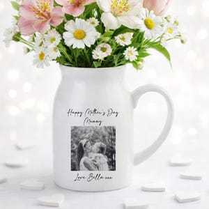 May include: A white ceramic mug with a black handle and the text "But First, Coffee" in a bold, black font. The mug is set against a blurred background. The mug appears to be approximately 10 cm tall.