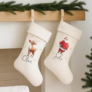 Può includere: Due calze natalizie color bianco sporco con illustrazioni ad acquerello di una renna e di Babbo Natale, personalizzate con i nomi Olive e Jole. Sopra, uno stendardo in tessuto recita Santa Please Stop Here for Olive & Jole con arte festiva.