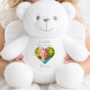 May include: A white plush teddy bear featuring angel wings on its back and a heart-shaped photo insert on its chest. The text above the heart reads A Cuddle From Heaven and the text below the heart reads Nanny, with a portrait of an elderly woman.