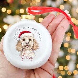 May include: White Christmas ornament with a watercolor illustration of a dog wearing a Santa hat. The ornament has the text "Have a Magical Christmas" and the dog's name, "Penny". A red ribbon is attached for hanging.
