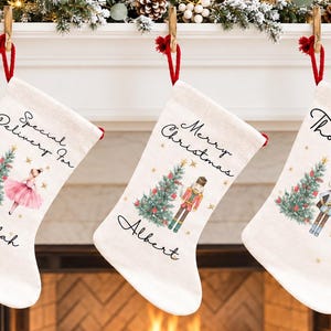 May include: Three off-white fabric Christmas stockings hanging from a mantel. Each stocking features a printed green Christmas tree, gold stars, and a unique character: a ballerina, a nutcracker, and a soldier. Text includes names and holiday phrases.