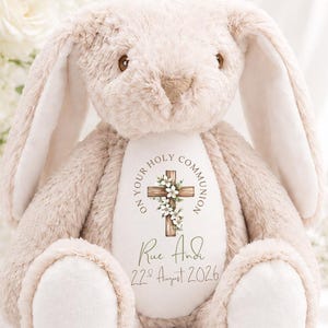 May include: A beige plush rabbit with long ears and a white belly patch. The belly features a printed wooden cross with white flowers, the text ON YOUR HOLY COMMUNION, the name Rue Andi, and the date 22nd August 2026 in a dark green font.