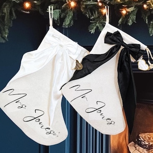 Peut inclure: Deux bas de Noël, un blanc et un blanc cassé, sont suspendus à une guirlande. Le bas blanc a un nœud blanc et les mots "Mrs Jones" en écriture noire. Le bas blanc cassé a un nœud noir et "Mr Jones" en écriture noire.