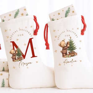 May include: Two off-white fabric Christmas stockings with red drawstring closures. Each features a teddy bear graphic, gold stars, and the text Special Delivery For. One stocking displays a large red letter M and the name Mason, while the other displays the name Amelia.