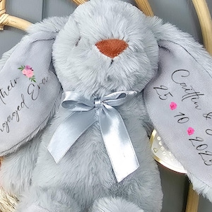 May include: A soft grey bunny toy with a silver ribbon and personalised text on its ears. One ear says "In their Engaged Era" with floral details. The other ear has the names "Caitlin & Sam" and the date "25.10.2025".