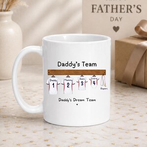 Op de afbeelding: Een witte keramische mok met een afbeelding van vier sportshirts die aan een houten rek hangen. De tekst luidt Daddy's Team en Daddy's Dream Team. De shirts zijn gelabeld met Daddy, Mummy, Rafe en Charlie en de nummers &eacute;&eacute;n tot en met vier.